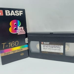 Puede incluir: Una cinta VHS BASF T-160 negra y su caja. La caja presenta el logotipo de BASF, un gráfico colorido de "8 hrs" y las palabras "Super High Grade". La cinta tiene una etiqueta manuscrita.