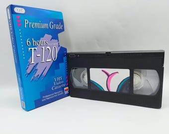 Może przedstawiać: Czarna kaseta wideo VHS obok niebieskiego kartonowego opakowania. Na opakowaniu znajduje się napis Premium Grade, 6 hours in EP mode, T-120 oraz VHS Video Cassette. Kaseta posiada przezroczyste okienko ukazujące wewnętrzne szpule.
