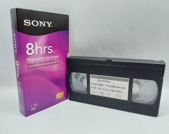 Pode incluir: Uma fita VHS Sony T-160 numa caixa roxa e preta rotulada 8hrs Premium, ao lado de uma cassete VHS preta com uma etiqueta branca escrita à mão que diz Blackstar, 13 Episodes - Complete Series, 4:41 - No Commercials e marca Sony.
