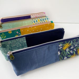 May include: A collection of colorful pencil cases with zipper closures. The cases feature various patterns, including floral, geometric, and solid color blocks. The zippers are in gold and silver tones. The cases are made of fabric.