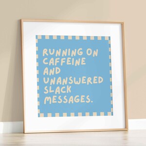 May include: A framed art print with the text "RUNNING ON CAFFEINE AND UNANSWERED SLACK MESSAGES." The artwork features a light blue background with a beige border and text in a handwritten style. The frame is a light wood color.