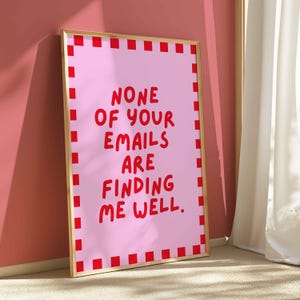 以下が含まれることがあります： 「NONE OF YOUR EMAILS ARE FINDING ME WELL」の赤い文字が入った、額入りのピンク色のアートプリント。プリントには赤い正方形のボーダーと金のフレームが付いています。アートワークは壁に立てかけられています。