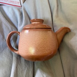 Puede incluir: Una pequeña tetera de cerámica con tapa, asa y boquilla. La tetera es de color marrón cálido moteado. El asa es curva y la boquilla es corta e inclinada. La tapa se asienta sobre la tetera.