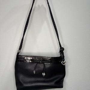 May include: Black leather shoulder bag with a braided strap. The bag features a drawstring closure with silver-toned accents and a heart-shaped charm. The bag has a textured leather body and a faux crocodile trim.