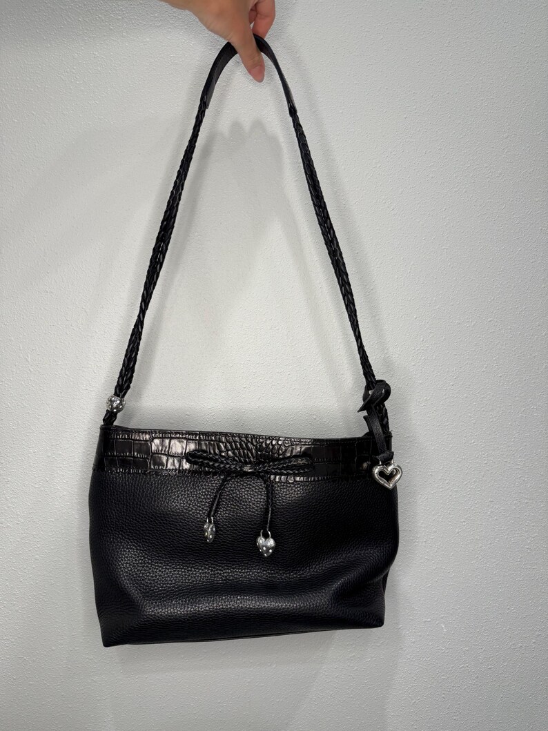 May include: Black leather shoulder bag with a braided strap. The bag features a drawstring closure with silver-toned accents and a heart-shaped charm. The bag has a textured leather body and a faux crocodile trim.