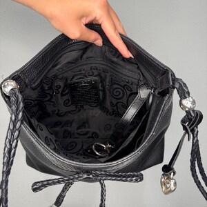 May include: Black leather crossbody bag with a braided strap and silver heart-shaped charms. The interior features a black paisley pattern. The bag is open, revealing the inside pocket and key ring.