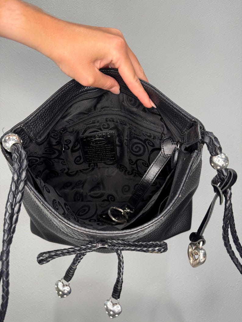 May include: Black leather crossbody bag with a braided strap and silver heart-shaped charms. The interior features a black paisley pattern. The bag is open, revealing the inside pocket and key ring.