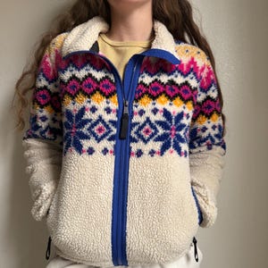 May include: A cream-colored fleece jacket with a vibrant geometric pattern in pink, blue, and yellow. The jacket features a blue zipper and collar, and two side pockets. The fleece material appears soft and warm, perfect for cooler weather.