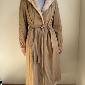 Puede incluir: Un abrigo color canela con capucha y cuello forrados de piel sintética. El abrigo tiene cinturón en la cintura, cierres de botones y mangas largas. El abrigo, de estilo vintage, llega a la rodilla y parece ser de material suave.