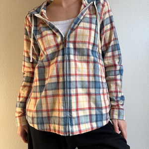 Könnte beinhalten: Ein karierter Kapuzenpullover mit Reißverschluss in Creme, Blau, Rot und Gelb. Der Hoodie hat eine Kapuze mit Kordelzug und einen durchgehenden Reißverschluss. Das Kleidungsstück ist aus einem weichen, möglicherweise Flanell-Material.