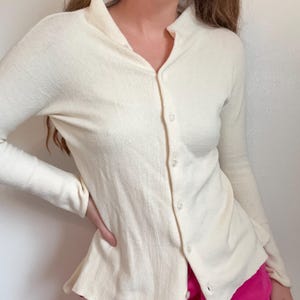 May include: Cream-colored button-down shirt with a high collar and long sleeves. The shirt is paired with bright pink sweatpants. The shirt has a textured appearance and is buttoned up the front.