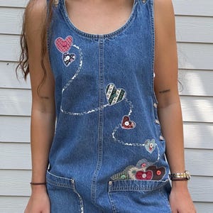 Può includere: Un vestito di jeans senza maniche con applicazioni decorative a forma di cuore in vari colori e motivi. L'abito presenta un top stile bavaglino, ampie tasche frontali e dettagli di cucitura bianchi. L'abito ha un'estetica casual e vintage.