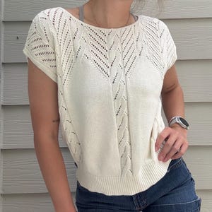 Pode incluir: Top de malha creme de manga curta com decote redondo. O top apresenta um design de malha de cabo na frente e trabalho aberto decorativo nos ombros. A bainha inferior e as mangas têm um acabamento canelado. O top é combinado com calções de ganga.