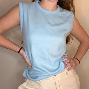 May include: A pale blue sleeveless jumper with a round neck. The jumper is worn with beige trousers. The outfit is casual and suitable for everyday wear. The jumper is made of a soft, knitted material.