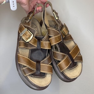 Può includere: Sandali in pelle marrone con più cinturini e chiusure con fibbia. I sandali hanno una suola spessa marrone scuro e cuciture a vista. Il marchio "Dr. Martens" è visibile sulla fibbia. Stile classico.
