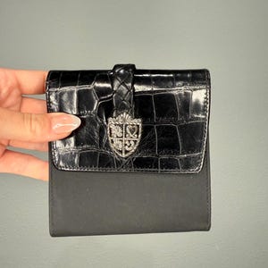 May include: Black wallet with a textured, crocodile-like flap and a smooth, matte body. The flap features a silver-toned emblem and a braided leather closure. The wallet is compact and appears to be made of leather.