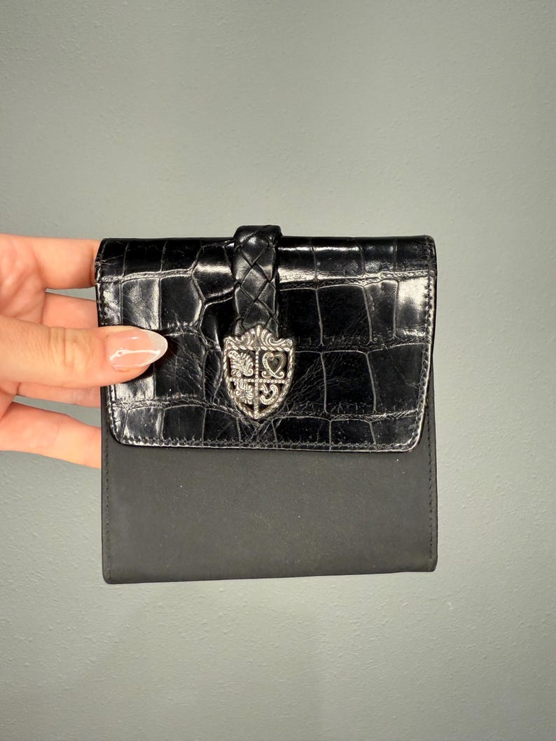 May include: Black wallet with a textured, crocodile-like flap and a smooth, matte body. The flap features a silver-toned emblem and a braided leather closure. The wallet is compact and appears to be made of leather.