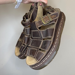 Può includere: Sandali in pelle marrone con punta aperta e cinturini multipli. I sandali hanno cuciture bianche e fibbie in ottone. Le suole sono spesse e marroni con una striscia bianca sul bordo.