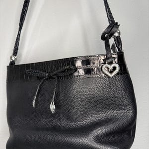 May include: Black leather shoulder bag with a textured finish and a faux crocodile trim. The bag features a braided strap, silver-tone hardware, and a heart-shaped charm. A drawstring closure with decorative silver-tone accents adds detail to the design.