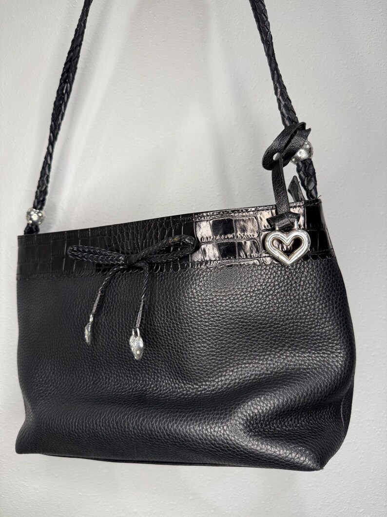 May include: Black leather shoulder bag with a textured finish and a faux crocodile trim. The bag features a braided strap, silver-tone hardware, and a heart-shaped charm. A drawstring closure with decorative silver-tone accents adds detail to the design.