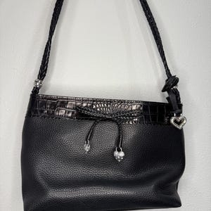May include: Black leather shoulder bag with a textured finish. The bag features a braided strap and a decorative top with a crocodile-like pattern. Silver-toned heart and jewel accents add detail to the design.