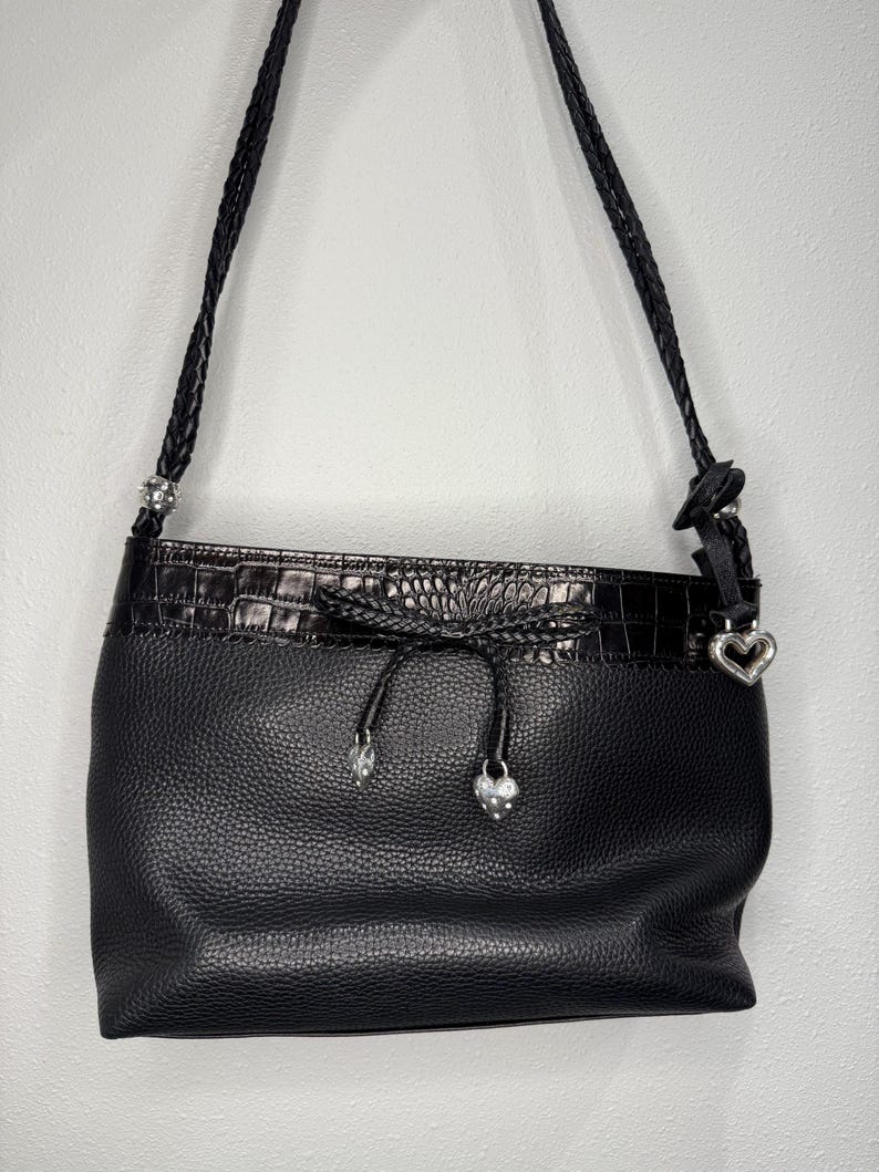 May include: Black leather shoulder bag with a textured finish. The bag features a braided strap and a decorative top with a crocodile-like pattern. Silver-toned heart and jewel accents add detail to the design.