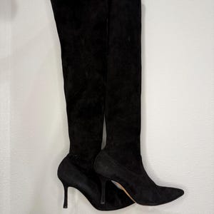 Puede incluir: Un par de botas negras por encima de la rodilla. Las botas están hechas de un material suave similar al ante y presentan una punta puntiaguda y un tacón de aguja. Las botas se sostienen contra un fondo blanco.