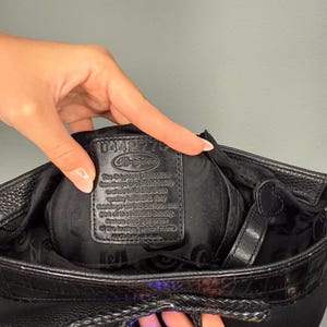 May include: Black leather Brighton handbag with a textured exterior. The bag features a braided drawstring closure and a decorative silver charm. The interior reveals a black lining and a leather tag with text.