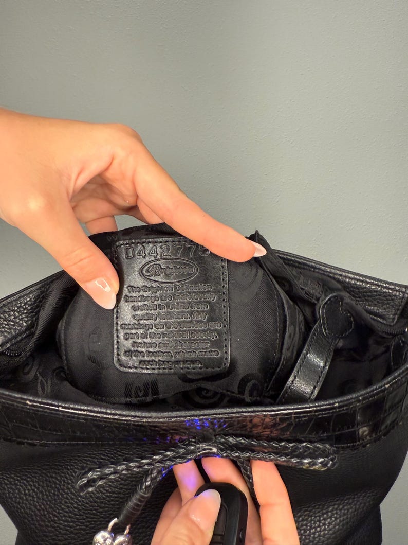 May include: Black leather Brighton handbag with a textured exterior. The bag features a braided drawstring closure and a decorative silver charm. The interior reveals a black lining and a leather tag with text.