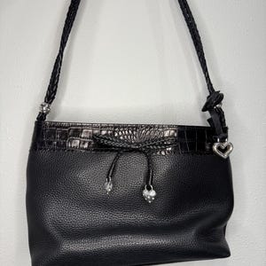 May include: Black leather shoulder bag with a textured finish. The bag features a braided strap, a decorative top with a crocodile pattern, and silver heart-shaped charms. The bag has a drawstring closure.