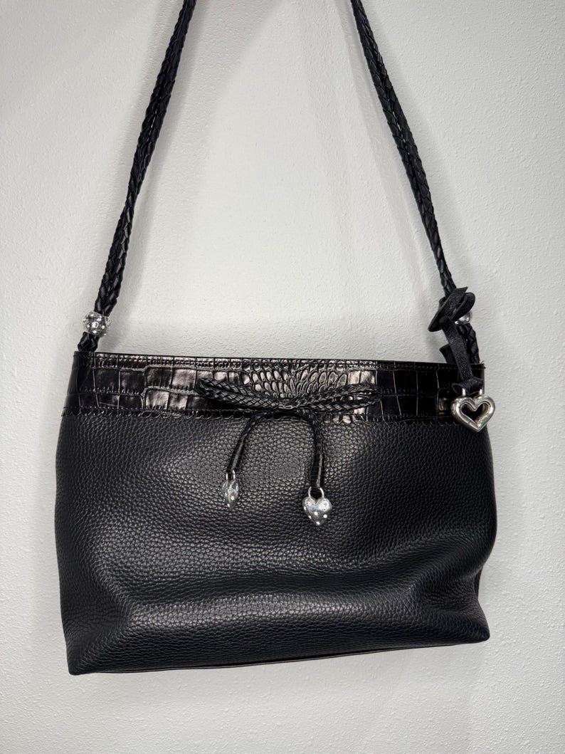 May include: Black leather shoulder bag with a textured finish. The bag features a braided strap, a decorative top with a crocodile pattern, and silver heart-shaped charms. The bag has a drawstring closure.