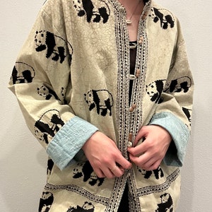 May include: A cream-colored button-up jacket with a black and white panda print. The jacket has a mandarin collar and a decorative beaded closure.