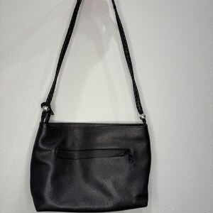 May include: Black leather shoulder bag with a braided strap. The bag has a textured surface and a zippered pocket on the back. The strap is held by a hand.