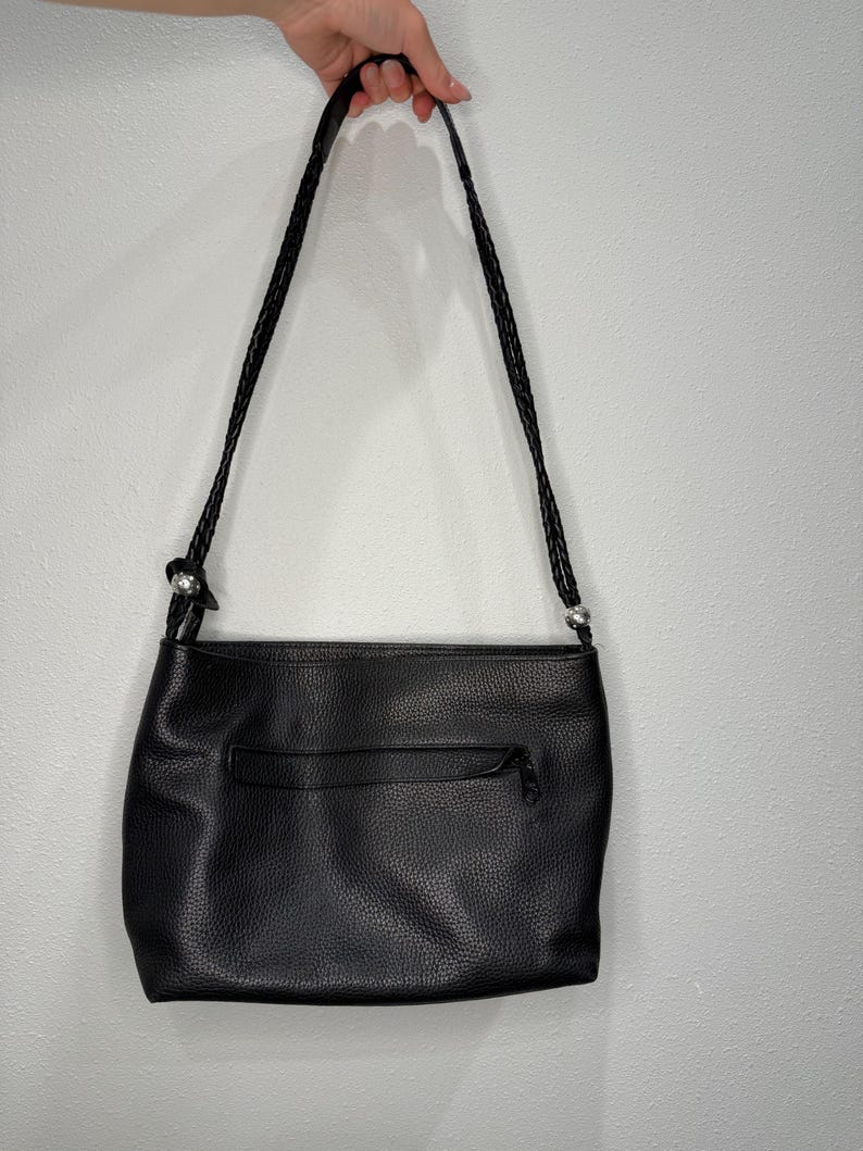 May include: Black leather shoulder bag with a braided strap. The bag has a textured surface and a zippered pocket on the back. The strap is held by a hand.