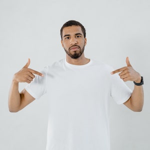 Könnte beinhalten: Ein Mann in einem weißen T-Shirt zeigt mit beiden Händen auf das Shirt. Er schaut direkt in die Kamera.