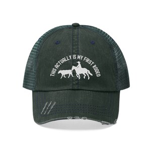 May include: A dark green and blue distressed trucker hat with a white embroidered design that says "This actually is my first rodeo" with a cowboy riding a horse and a calf.