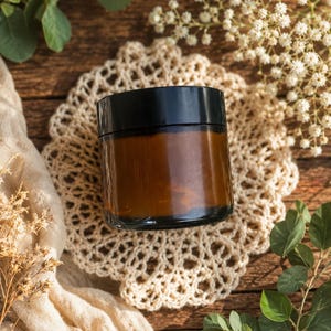 Op de afbeelding: Een bruine glazen pot met een zwarte deksel staat op een crèmekleurig gehaakt kleedje. De pot is omgeven door groen en kleine witte bloemen, alles gerangschikt op een houten oppervlak. De pot lijkt een product te bevatten.