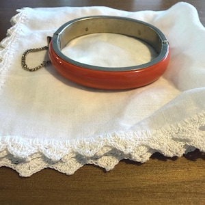 May include: A hinged bangle bracelet featuring a smooth, vibrant orange exterior and a metallic silver-toned interior. The bracelet includes a small, attached safety chain with a clasp mechanism, providing a secure closure for the circular band.