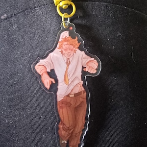 May include: An acrylic keychain featuring a cartoon illustration of a person with messy orange hair, wearing a white button-down shirt with a loose yellow tie and brown trousers. The figure is in a dynamic pose, attached to a yellow lobster clasp.