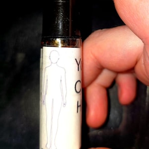 May include: A small, cylindrical glass bottle with a black cap. The label features a line drawing of a human figure and the letters "YCH". The bottle is held against a dark background.
