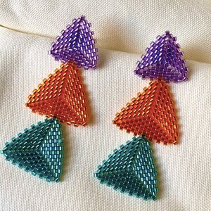 Può includere: Un paio di orecchini pendenti con tre perline triangolari. La perlina superiore è viola, la perlina centrale è arancione e la perlina inferiore è turchese.