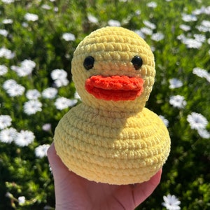May include: A yellow crocheted duck toy with red felt beak and black button eyes. The duck is being held in a hand against a background of white flowers.
