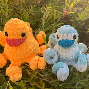 May include: Two crocheted octopus plushies, one orange and one blue and white. The orange octopus has a pink beak and black eyes. The blue and white octopus has a blue beak and black eyes. Both are sitting on a bed of green grass.