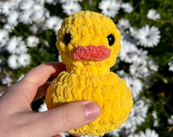 May include: A yellow crochet duck with black eyes and a pink beak. The plush toy is held in a hand against a backdrop of white flowers and green foliage. The duck is a soft, textured item, likely handmade.
