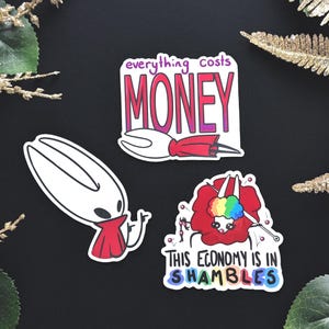 以下が含まれることがあります： 黒い背景に3つのステッカー。1つは紫とピンクで「everything costs MONEY」と書かれています。もう1つは赤いマントを着た白いキャラクターです。3つ目は、虹色の毛糸玉で「THIS ECONOMY IS IN SHAMBLES」と書かれています。