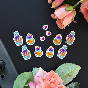 May include: Nine colorful stickers featuring small glass bottles with purple ribbons and orange liquid inside. The stickers are arranged in a semi-circle on a black surface. There are also four small pink heart stickers on the surface.