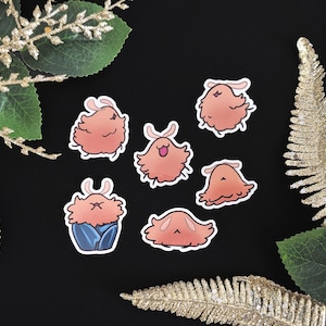 May include: A collection of six stickers featuring cartoonish, fluffy creatures in shades of peach and pink. Some have small, curved antennae. One sticker shows the creature in a blue outfit. The stickers are arranged on a black background, with gold and green decorative elements.