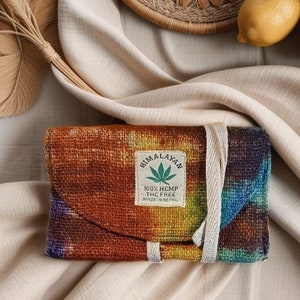 May include: A colorful, rectangular hemp wallet with a flap closure and a cream-colored tie. The wallet features a vibrant tie-dye pattern in shades of orange, yellow, blue, and purple. A label reads "Himalayan 100% Hemp THC Free Made in Nepal."