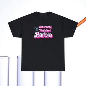 Op de afbeelding: Zwart T-shirt met een roze afbeelding met de tekst "Laboratory Assistant Barbie" met een gestileerde blauwe en witte vlinder.