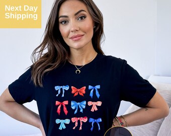 May include: A black short-sleeve t-shirt featuring a graphic print of nine decorative bows in various shades of red, white, and blue. The bows are arranged in a grid pattern on the front of the shirt, showcasing different patterns including stripes, polka dots, and solid colors.